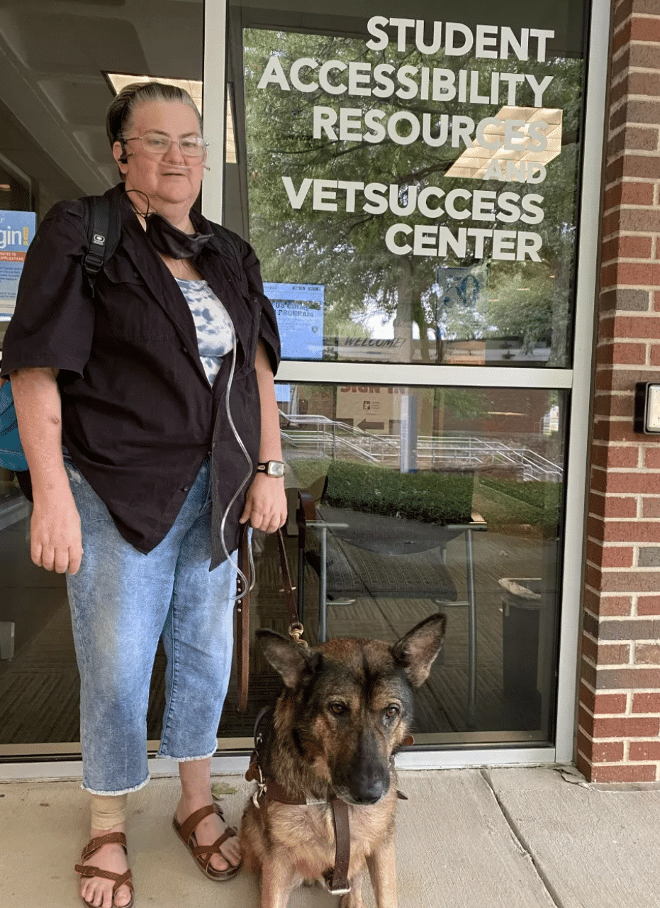 Jamie stands holding Haddie's harness as she sits in front of the resource center.