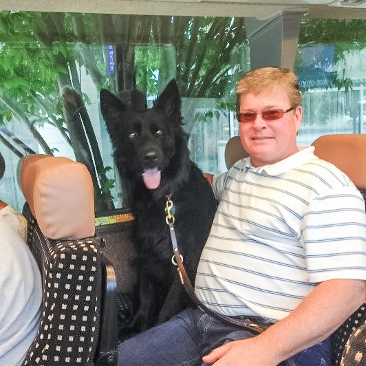 gary sitting on a bus seat next to his black guide dog rueben who has his mouth open and tongue out