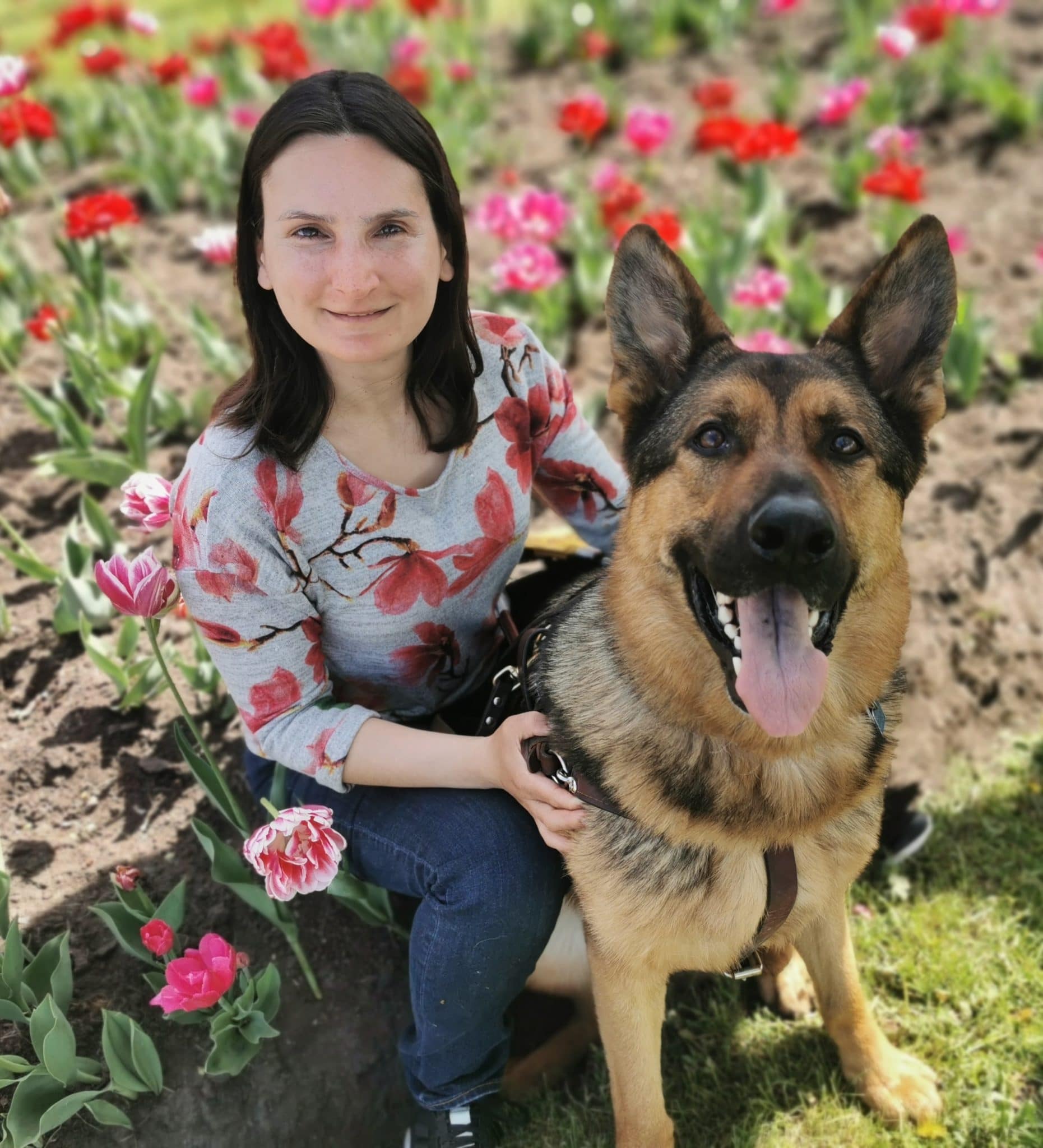 Esther sits in a field of flowers holding the harness of Zion her german shepherd guide dog in harness, both are smiling up at the camera